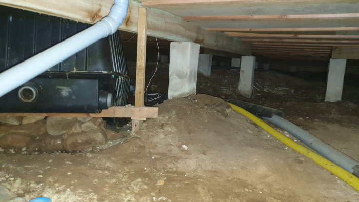 Utrymme under hus med oanvänd svart tank, rörledningar och stödben i betong.