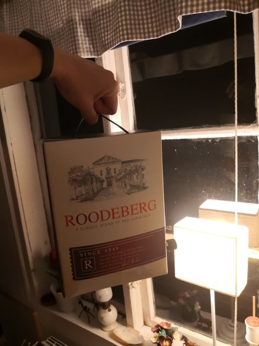 Hand håller en dunk vin bredvid ett fönster i ett mysigt inrett rum.