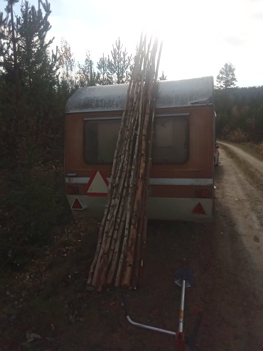 Björkved staplad mot husvagn med yxa och klyvyxa i förgrunden omgiven av skog.