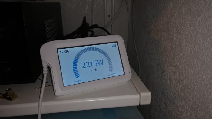 Sparsnäs energimätartillbehör på vit hylla som visar aktuell energianvändning på 2215W mot väggbakgrund.