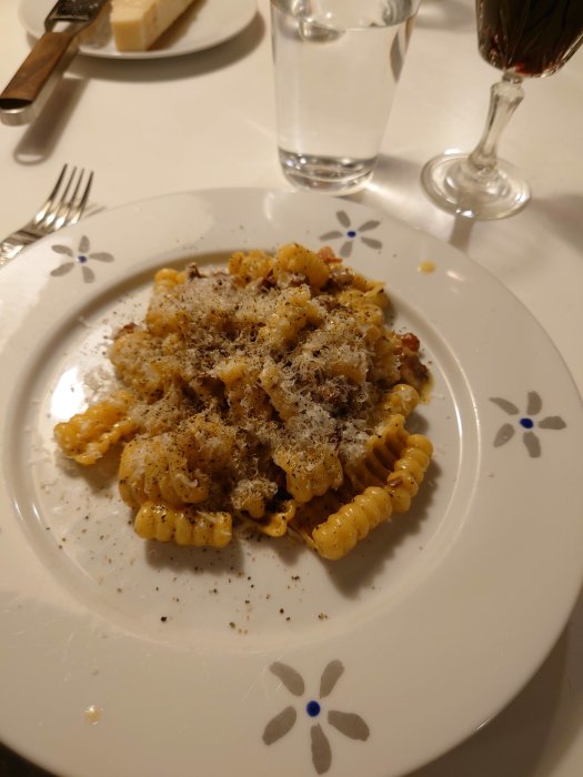 Tallrik med pasta carbonara toppad med riven parmesan, svartpeppar, bredvid ett glas vatten och vin.