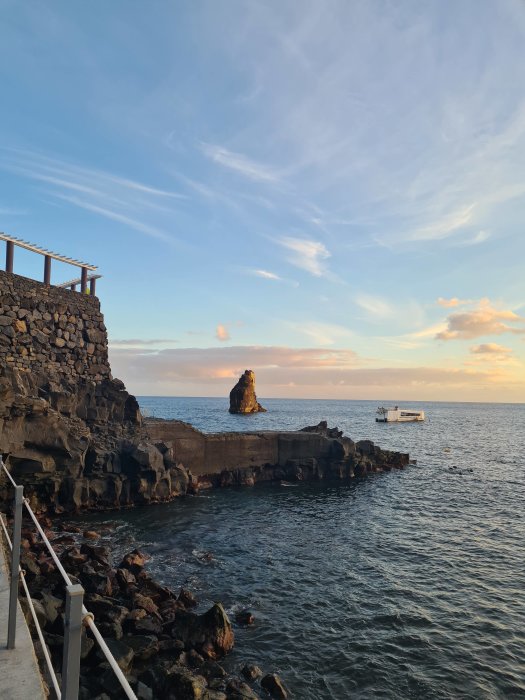 Solnedgång vid havet med en klippformation i vattnet och en båt i fjärran, stenmur och trappa i förgrunden.