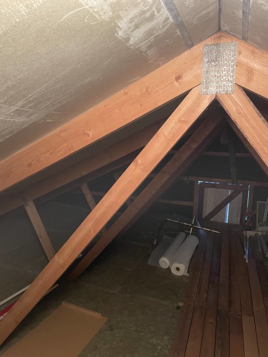 Inre delen av ett vindstak med synliga träbjälkar och en rullad isoleringsmatta på golvet.