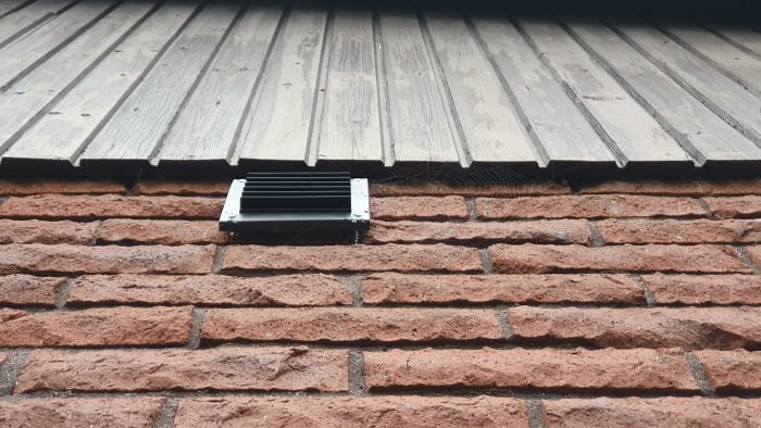 Plastventilationsgaller som inte passar jämnt mot ojämnt tegelvägg under ett träfärgat takutsprång.