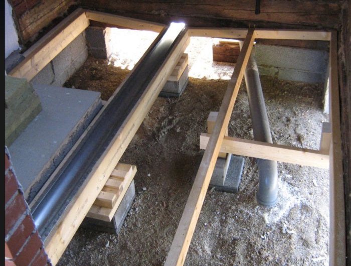 Byggkonstruktion med träbjälklag, ränna i metall och synligt avloppsrör, under arbete i ett renoveringsprojekt.