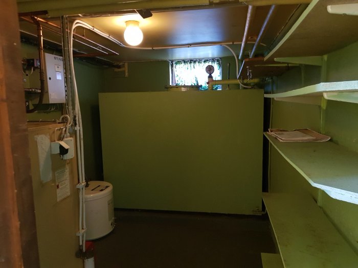 En källare med rördragning över en varmvattenberedare och en grön oljecistern.