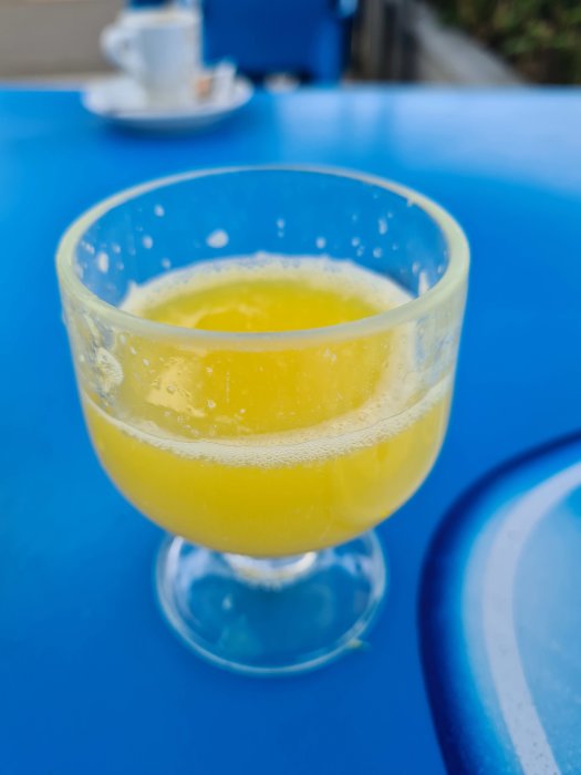 Ett glas med apelsinjuice på ett blått bord, suddig vit kaffekopp i bakgrunden.