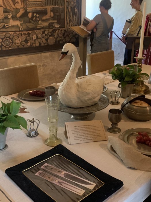 Dukat bord med en dekorativ svanfigur, tallrikar, bestick och besökare i bakgrunden som betraktar en väggbonad.