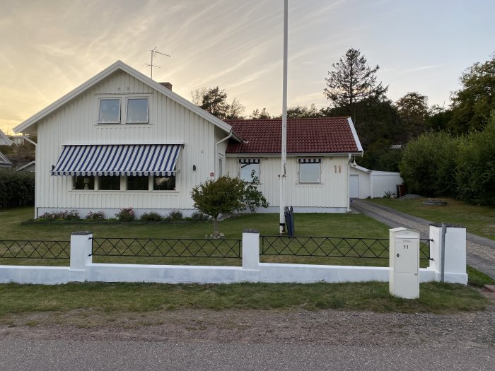 Villa med vit träfasad och rött tegeltak, randig markis och vitt staket.