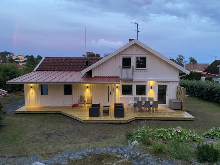 Villa med äggskalvit träfasad och rött tegeltak i skymningen, belysning på veranda, och ännu ej färdigmålat staket.