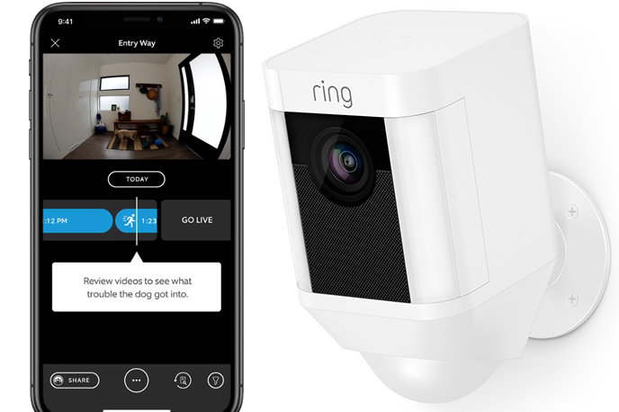 En vit Ring Spotlight Cam Battery bredvid en smartphone som visar appens gränssnitt med live video från kameran.