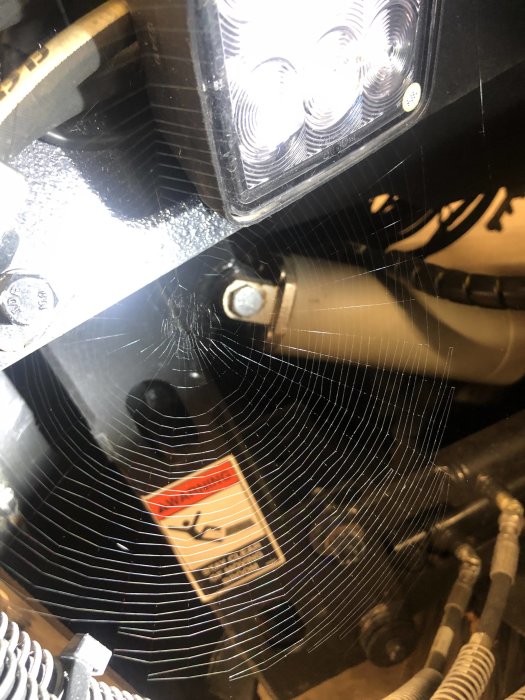 Närbild på fordonsdel med LED-ljus och spindelnät, varningsetikett synlig.