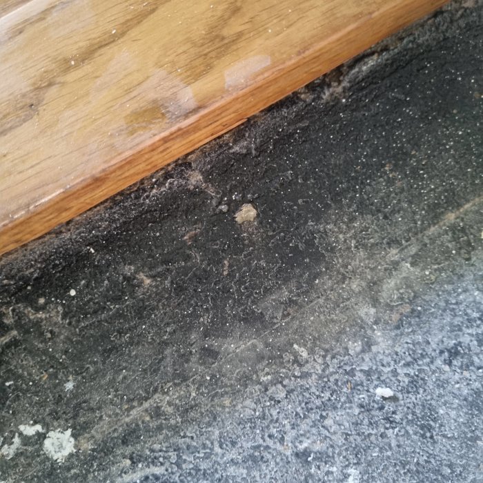 Ett gammalt golvlager under uppriven laminatklinker och board, visar svart materia möjligen limrester eller underlagsmatta, vid en trälist.