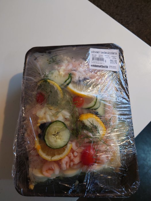 Förpackad smörgåstårta med räkor, citron, dill och gurka under plastfolie.