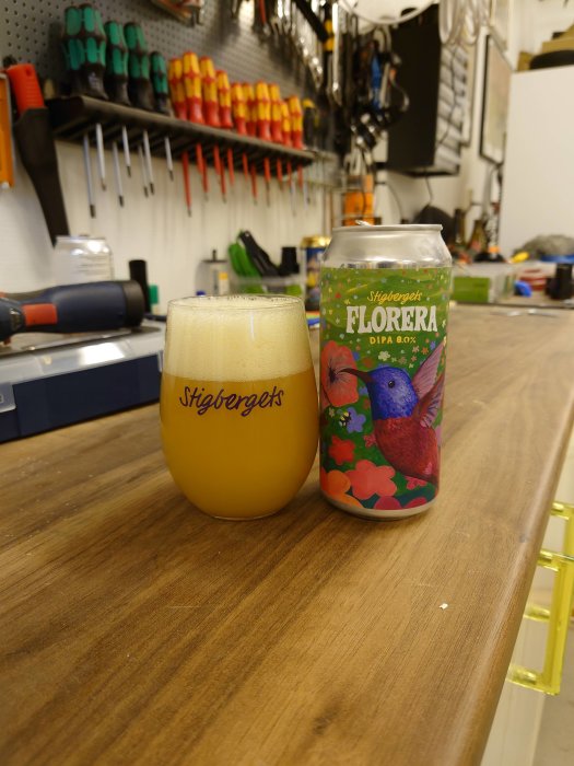 Ölglas och burk av Stigbergets FLORERA-öl på ett arbetsbord med verktyg i bakgrunden.