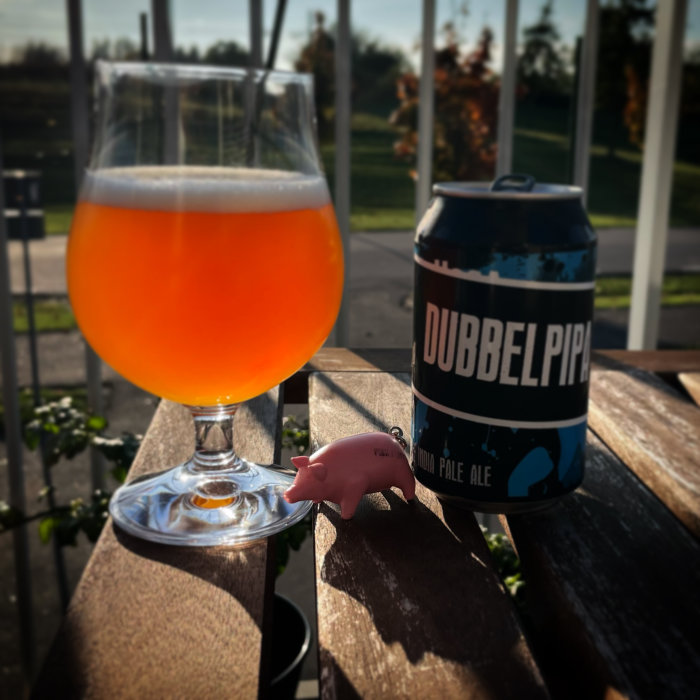 Ett glas öl på ett bord bredvid en burk Dubbelpip India Pale Ale och en liten plastfigur av en rosa gris.