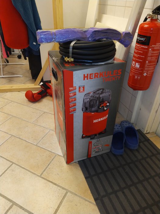 Ny Herkules Twenty-kompressor i kartong med tillbehör på golvet, bredvid brandsläckare och blåa skor.