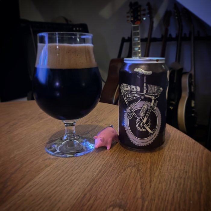 Ett glas mörkt öl och en ölburk med motorcykeldesign, en liten rosa grisfigur och gitarrer i bakgrunden.