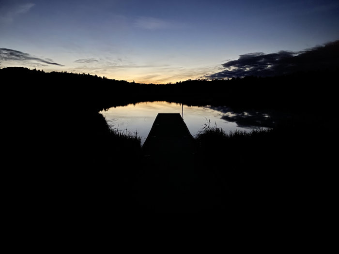 Brygga som sträcker sig ut i en stilla sjö vid skymning med spegelbild av himlen och träd i vattnet.