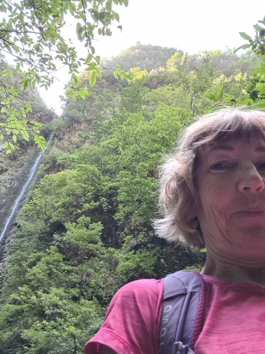 Kvinna tar selfie med ett vattenfall och grönskande skog i bakgrunden under en vandringspaus.