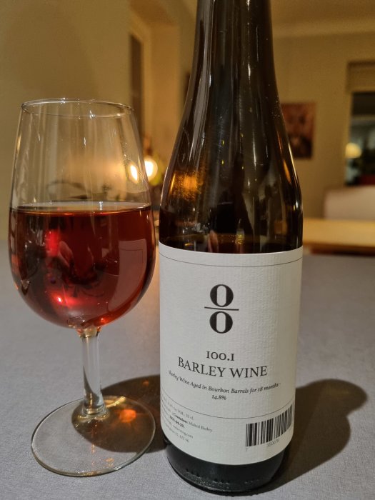 Ett glas fyllt med rödbrun barley wine bredvid en flaska på ett bord.