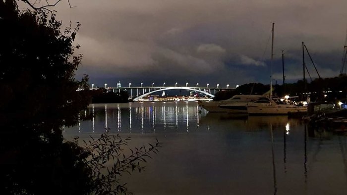 Kvällsutsikt över en upplyst bro och båtar i en småbåtshamn med mörka moln i bakgrunden.