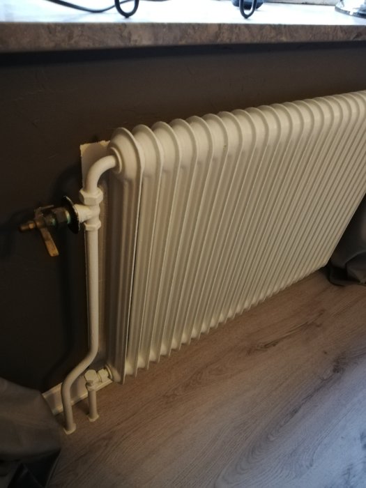 Vit gammal radiator med oanvändbara termostatvred synlig vid ett fönster, indikerar behov av underhåll.