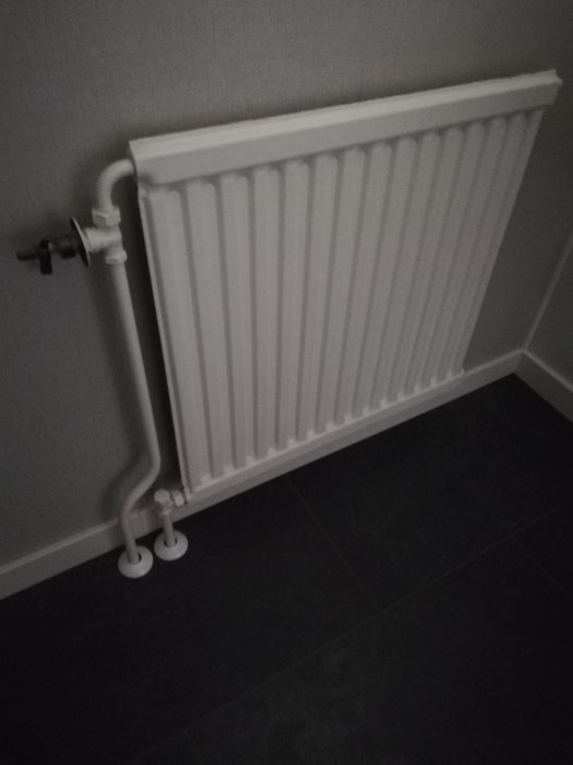 Gammal vit radiator med oanvändbar termostat monterad på en mörkgrå vägg.