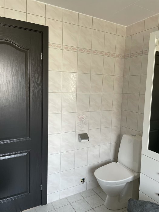 Renoverat badrum med nyspacklade vita kakelväggar, dekorationslist och svart dörr.