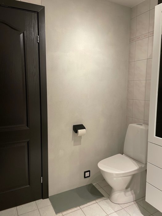 Renoverat badrum med nytt spacklat klinkergolv och delvis kaklade väggar, toalett och mörk dörr.