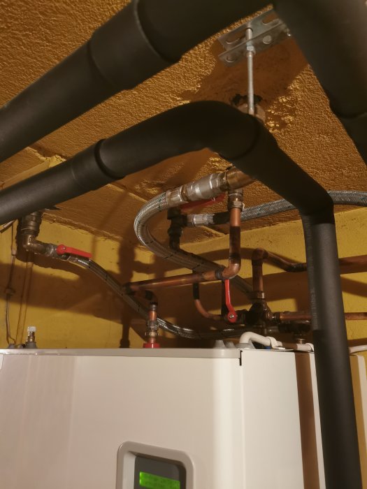 Värmesystem med kopparledningar och isolerade rör ovanför en vit värmepanna i ett rum med gula väggar.