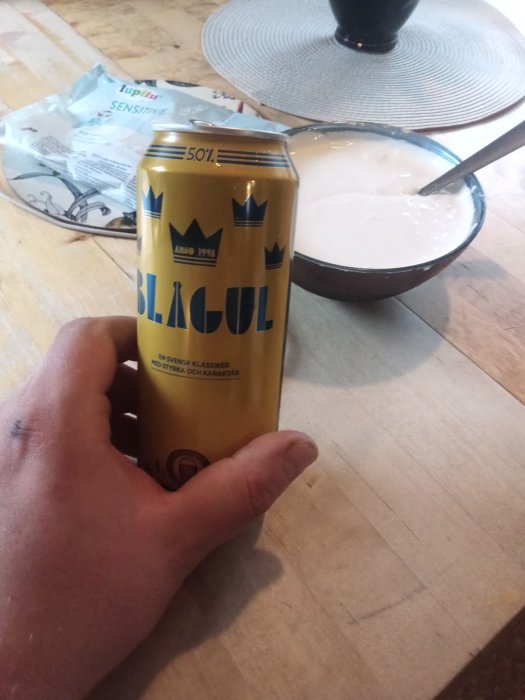 Hand håller en burk Blågul öl med en skål tzatziki och förpackningar i bakgrunden.
