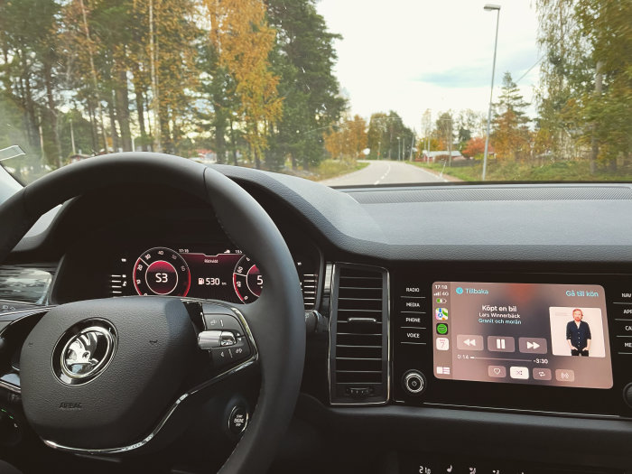 Bilinteriör med ratten i förgrunden och en landsväg synlig genom framrutan, multimedia-skärm visar en podcast.