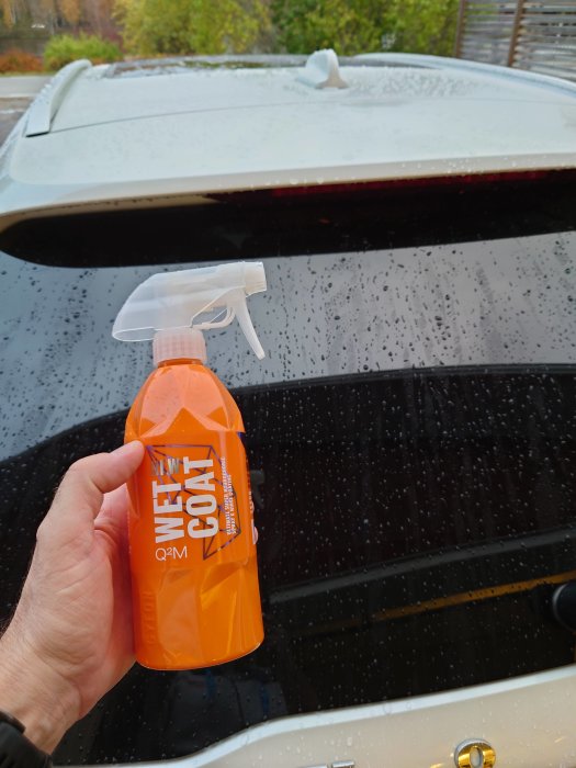Hand håller en orange sprayflaska med texten "WET COAT" framför en bil i regnet.