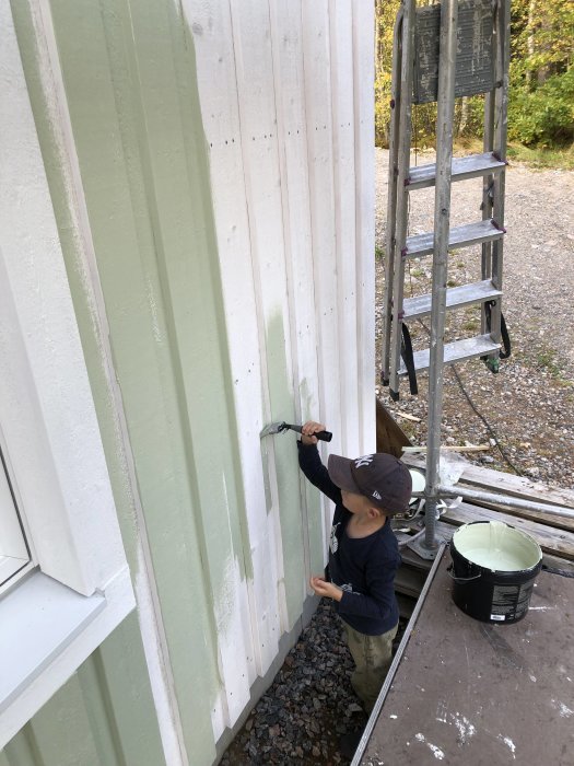 Barn hjälper till att måla husets träfasad med grön färg, stående nära en stege och färgburk.