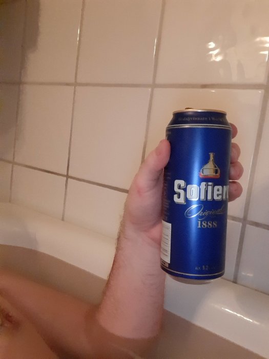 Hand håller en ölburk med märket Sofiero vid ett badkar med kakelvägg i bakgrunden.