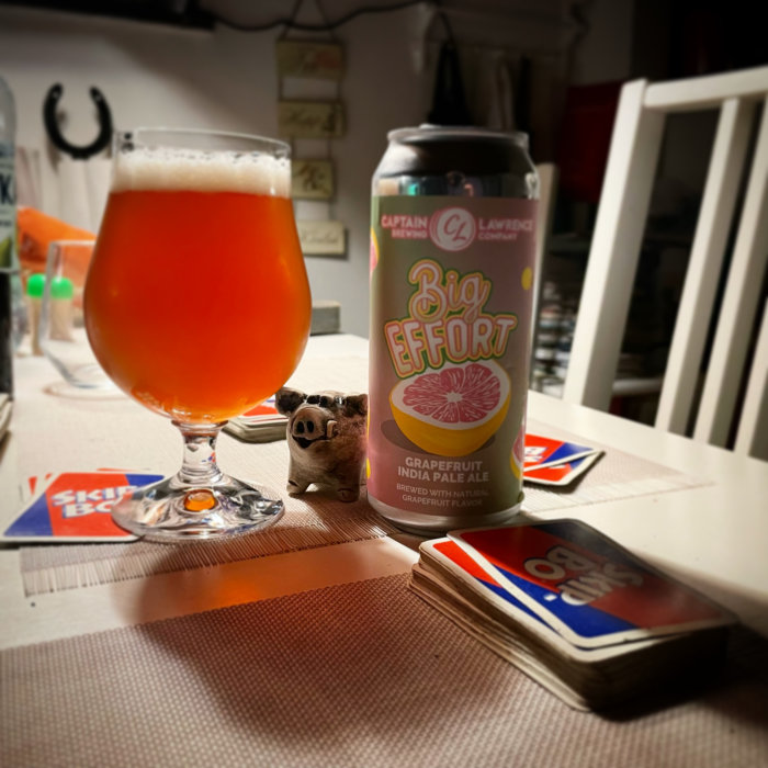 Ett glas öl och en burk IPA med namnet "Big Effort" bredvid en liten grisfigur och en kortlek på ett bord.
