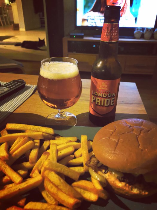 Hamburgare med pommes frites och en flaska London Pride öl på ett bord framför en TV.