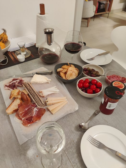 Charkbricka med skinka, salami, ost och tillbehör som oliver, tomater, chips, två fyllda vinglas och karaff.