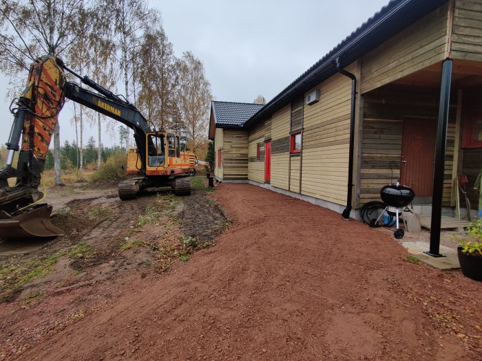 Grävmaskin bredvid hus under renovering med nygrusad yta framför till blivande entré/uteplats.