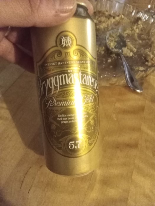 En hand håller en guldfärgad ölburk märkt "Bryggmästarens Premium Gold" med en delvis synlig maträtt i bakgrunden.