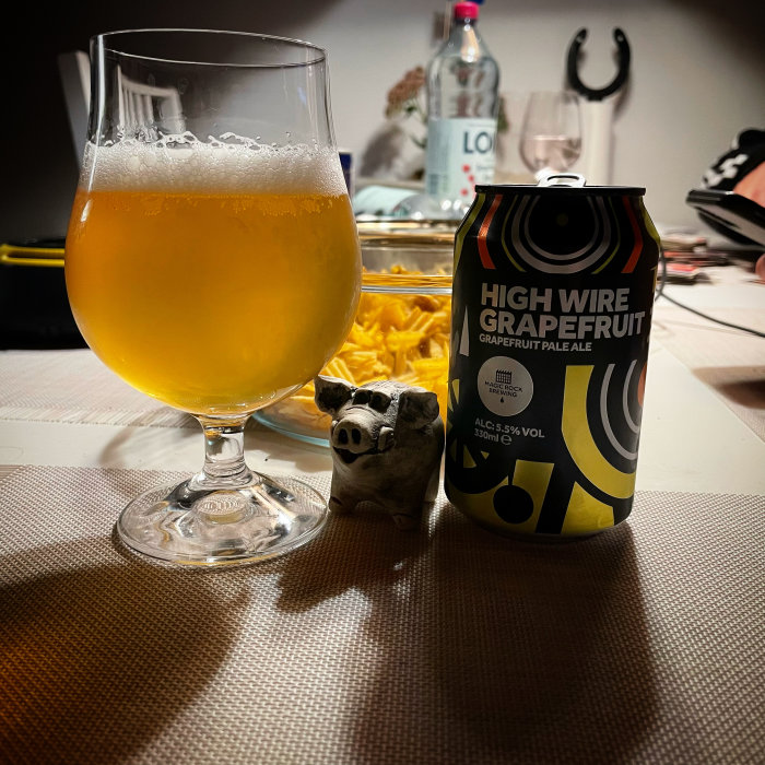 Glas med ljusgul öl nära en High Wire Grapefruit burk och en figur av en gris, med snacks och flaskor i bakgrunden.