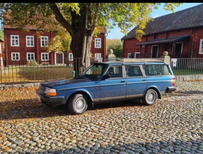 Blå Volvo 240 Classic -93 parkerad på kullersten framför röda trähus.