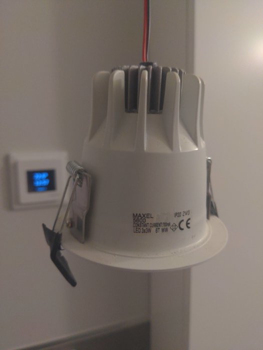 Nedpendlad LED-spotlight Maxel Sirius med tekniska specifikationer synliga, hängande vid en vägg med dimmerströmbrytare i bakgrunden.
