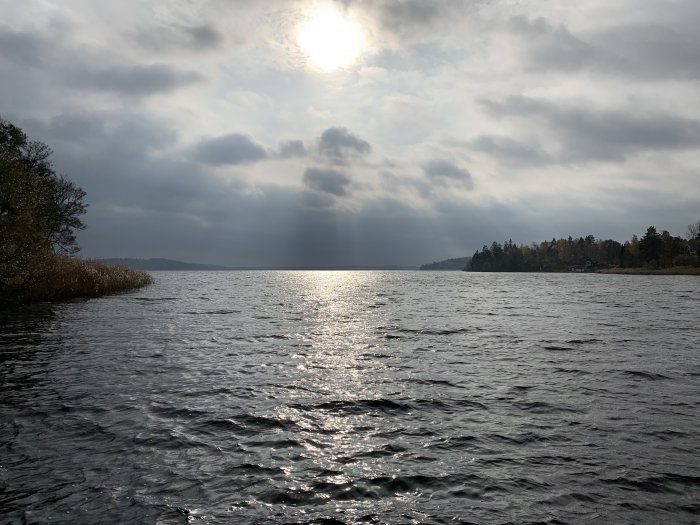 Sjövy med solen som speglar sig i vattnet, omgiven av moln och skogklädda kuster.