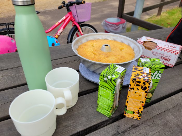Fika utomhus med saft, kakor, en kaka i form och två koppar på ett bord, med en rosa cykel i bakgrunden.