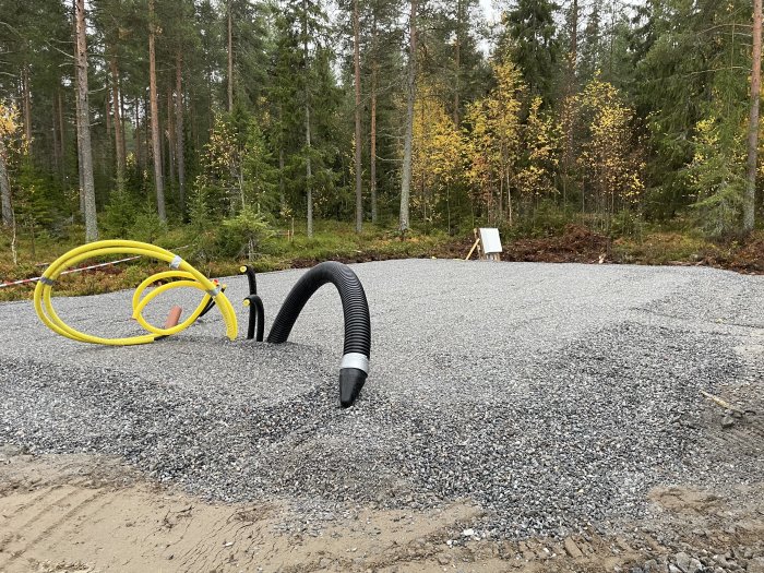 Nyanslutna avlopps-, vatten- och elkablar med kantbalk och isolering på grusunderlag vid skogstomt.