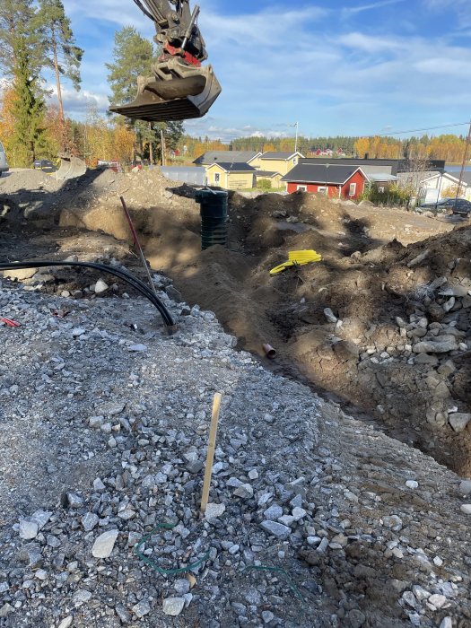 Grävmaskinskopa ovanför en grävd schaktgrop med avloppsrör, kablar och första isoleringslagret vid byggarbetsplats.