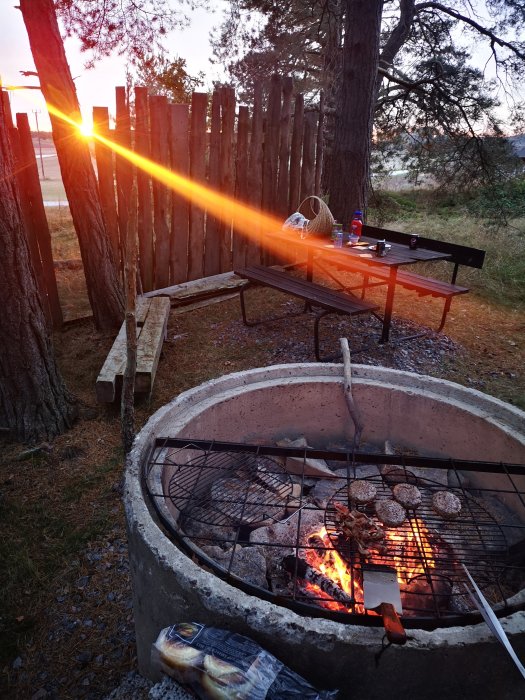 Grillning utomhus med hamburgare på grillen och solnedgång i bakgrunden.