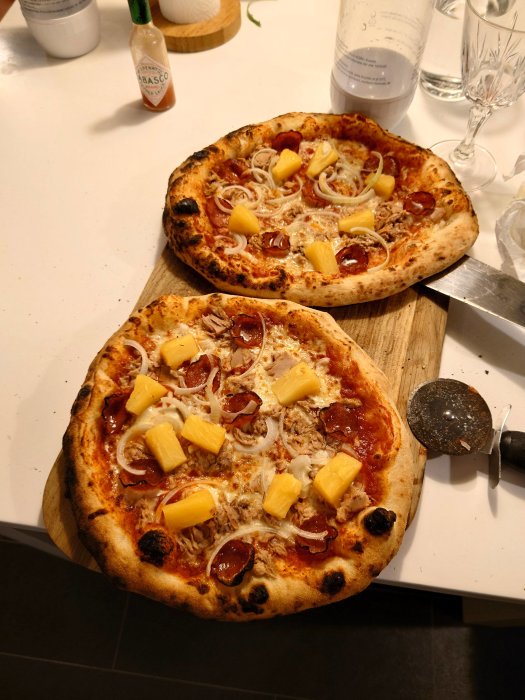 Två hemlagade pizzor med tonfisk, salami, lök, ananas och tabasco på ett köksbord.